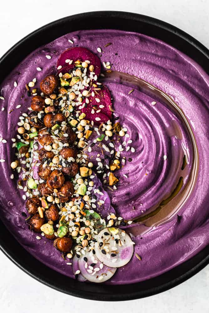 purple carrot hummus in a black bowl topped with colorful radishes, nuts, crispy chickpeas, seeds and spices; overhead shot on a white background