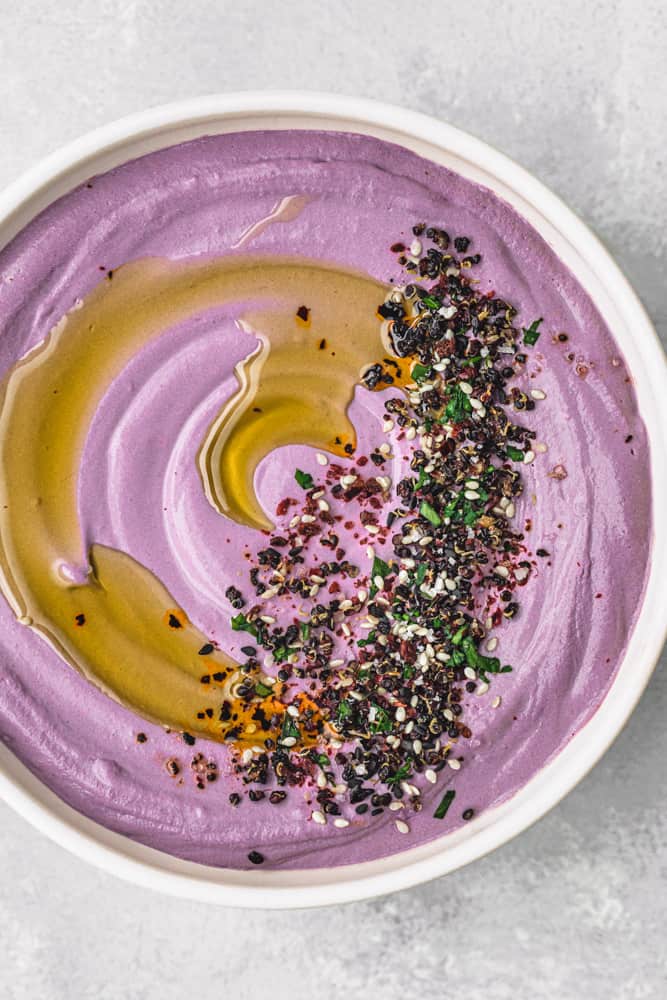 purple carrot hummus topped with olive oil, herbs, seeds and spices in a white bowl; overhead shotI