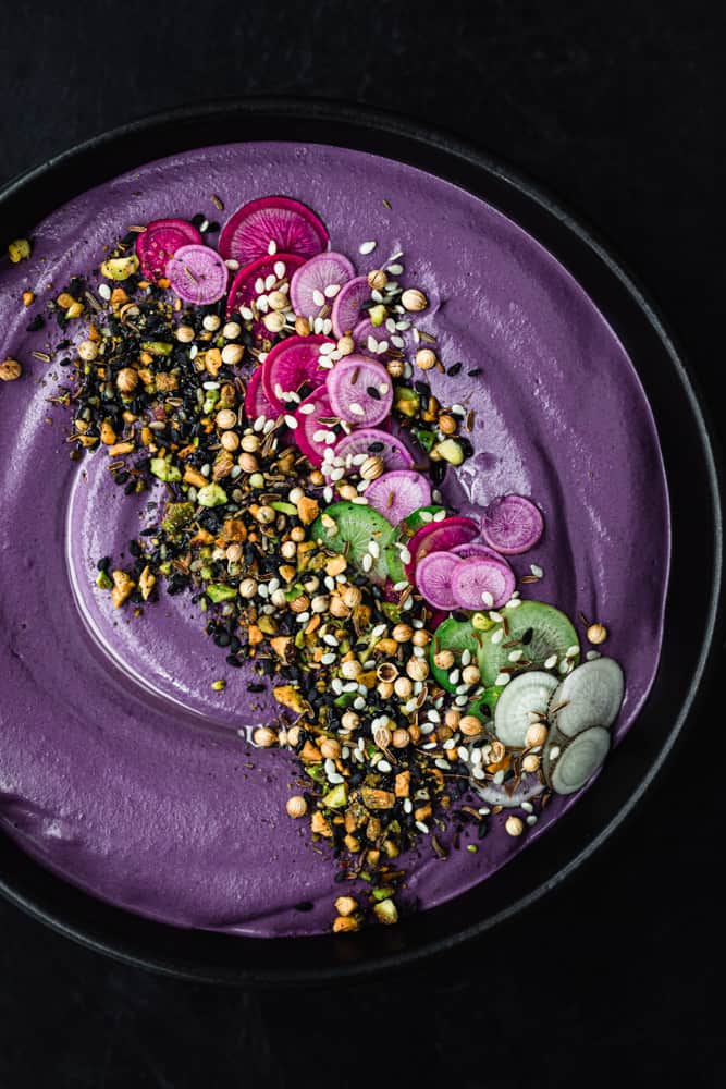purple carrot hummus topped with pink, purple and green thinly sliced radishes, nuts and seeds; in a black bowl on a dark background; overhead shot