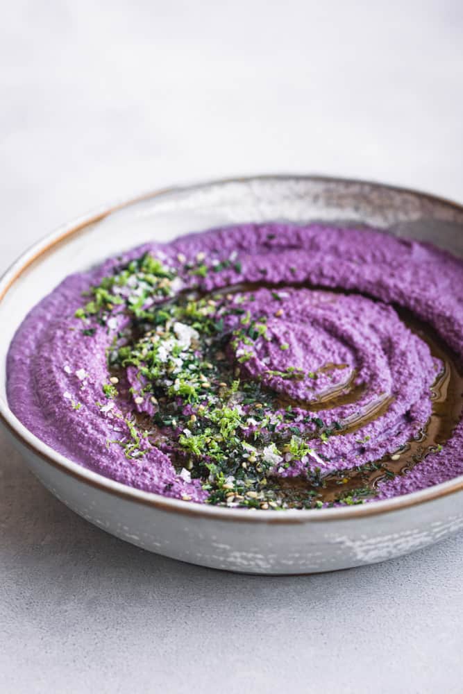 purple carrot hummus topped with lemon zest, fresh herbs and olive oil; side angle shot on a light background