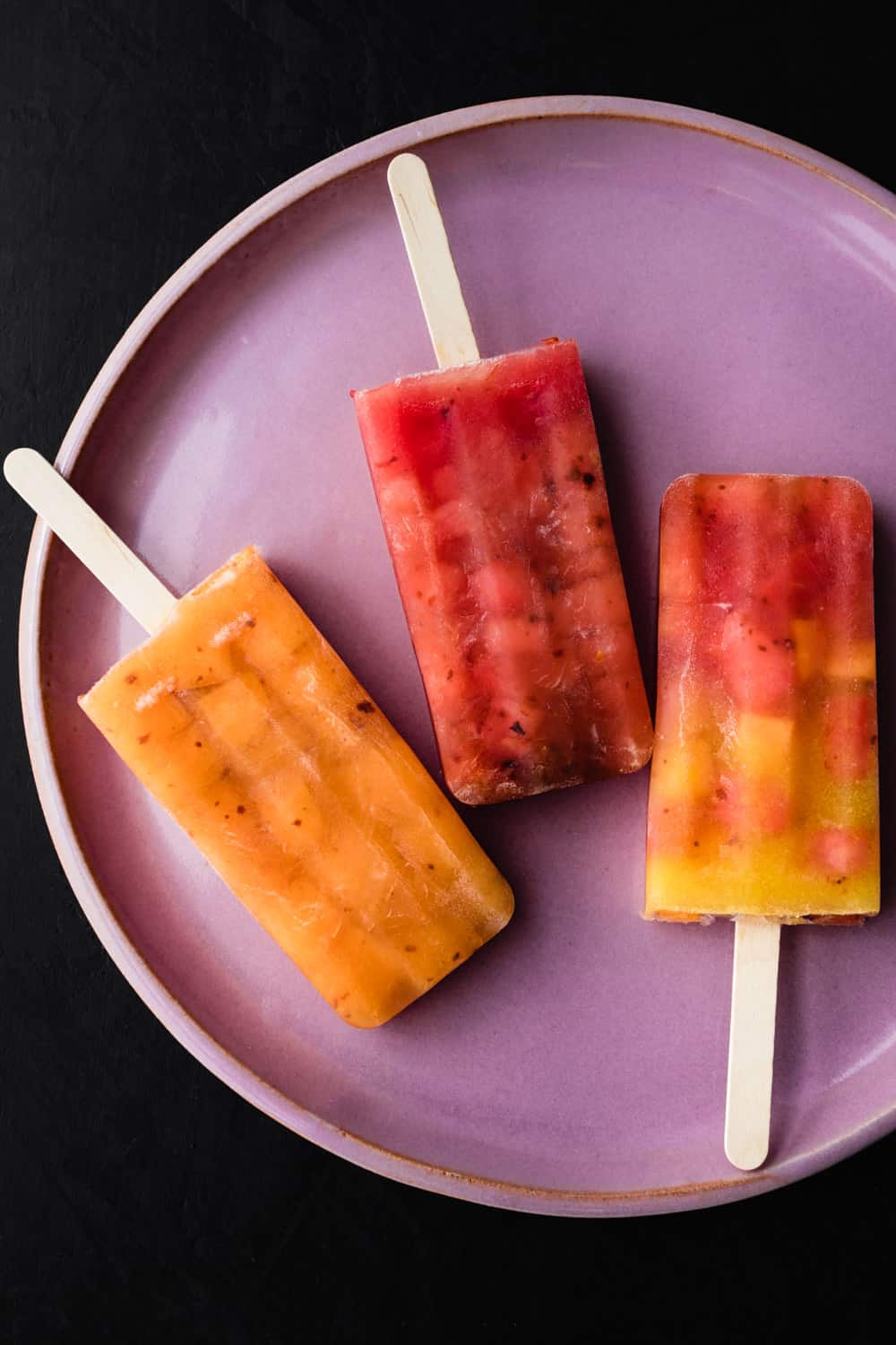 3 colorful watermelon popsicles on a pink plate; overhead shot on a black background