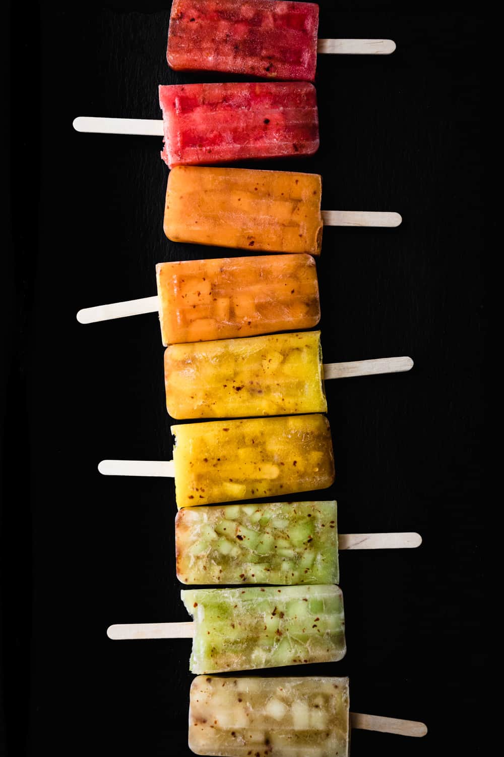 a popsicles rainbow! Red, pink, orange, yellow, green and white melon and chile popsicles lined like a rainbow; overhead shot on a black background