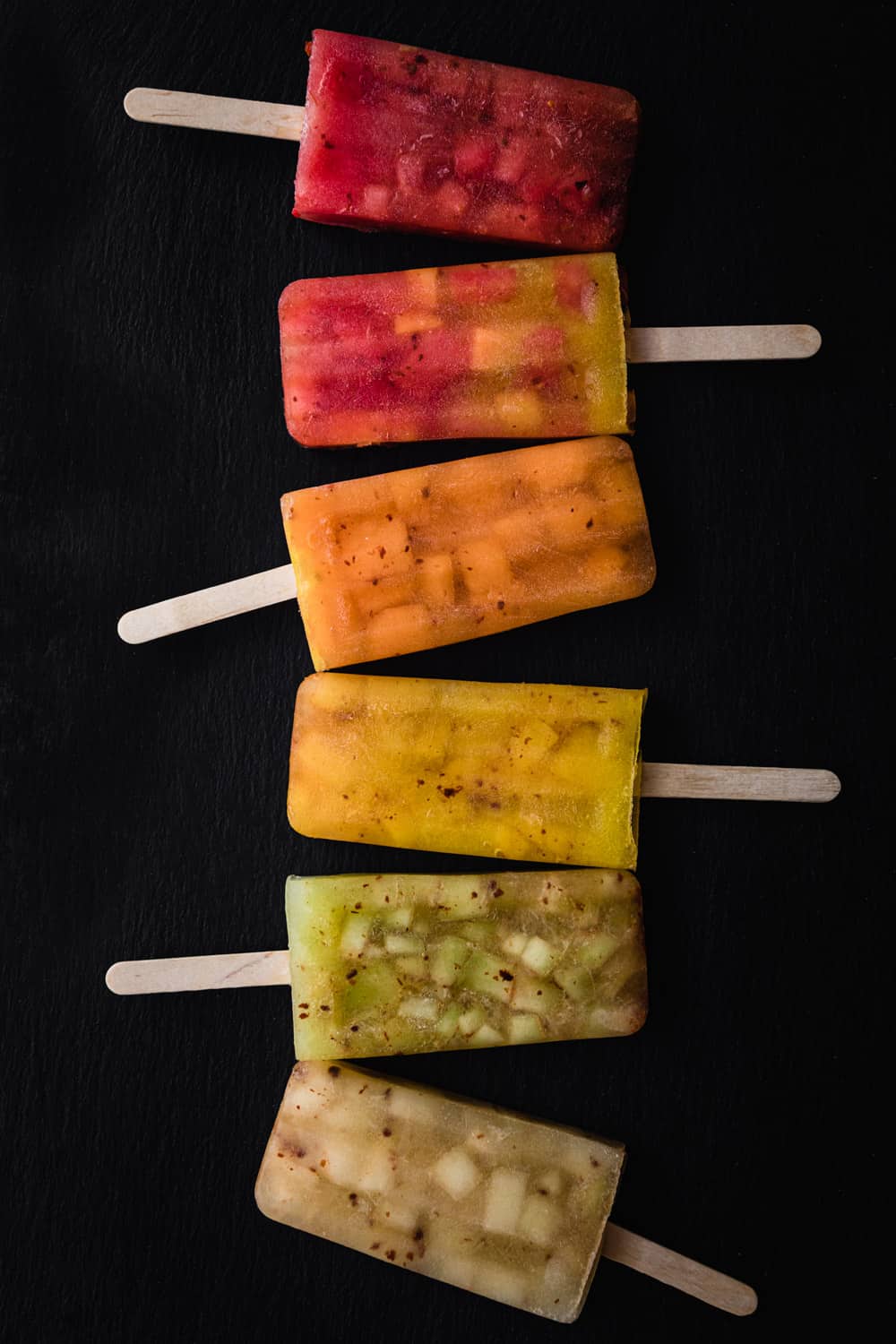 5 melon popsicles in red, orange, yellow, green white and mixed colors lined up on a black background; overhead shot