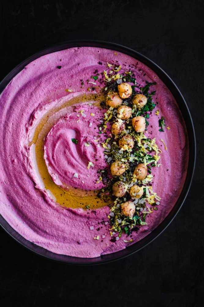 purple carrot hummus topped with garbanzo beans, zest, fresh herbs, spices and olive oil; in a black bowl on a black background