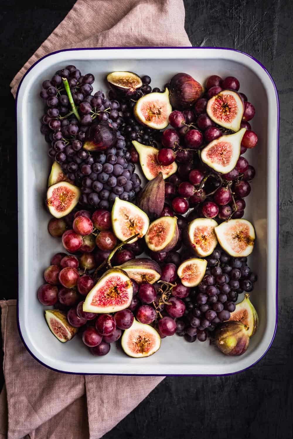 grapes and figs in a sheet pan on top of a pink linen; overhead on a dark backdrop