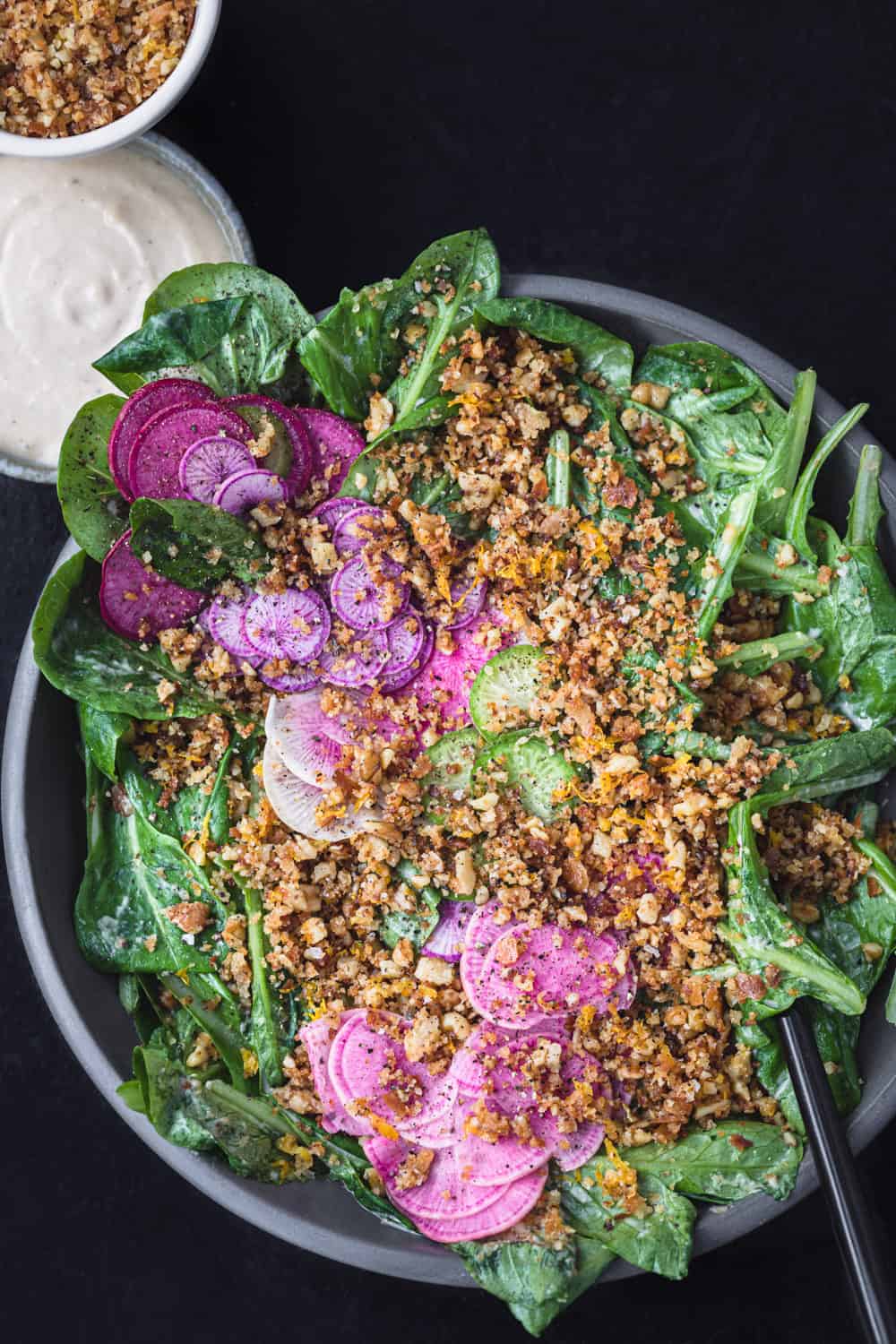A tahini caesar salad in a grey bowl with lots of breadcrumbs and radishes; extra dressing and breadcrumbs in little bowls on the side; overhead shot