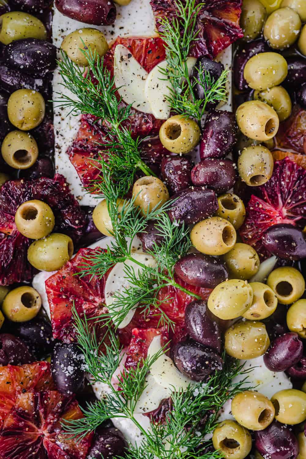 Feta topped with olives, blood oranges and fresh dill; in a baking dish and overhead shot.