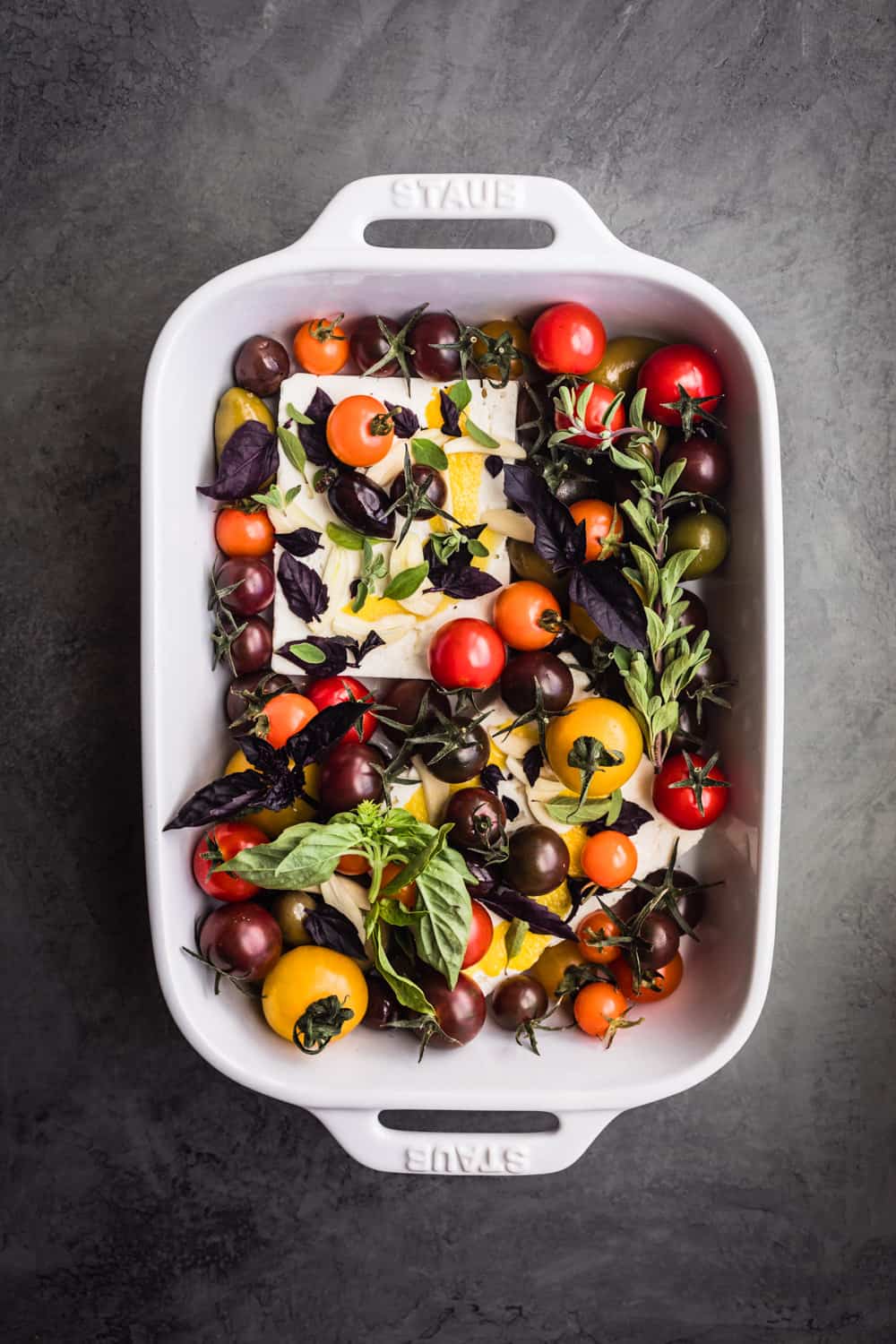 Feta in a baking dish with tomatoes, fresh herbs, lemon zest and garlic; overhead shot.