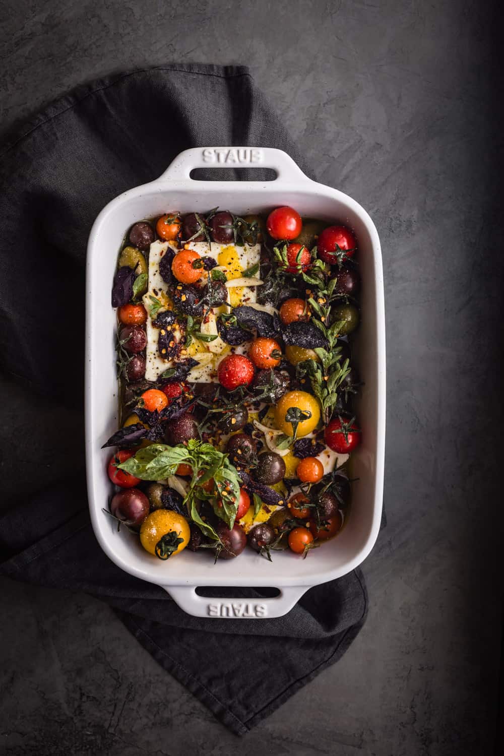 Feta with tomatoes, garlic, lemon, and fresh herbs in baking dish; overhead shot.