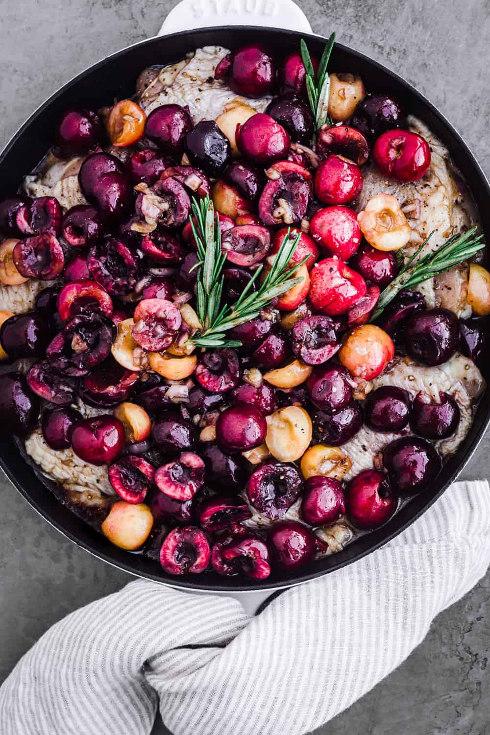 Raw chicken thighs topped with cherries and rosemary in a skillet; overhead shot.