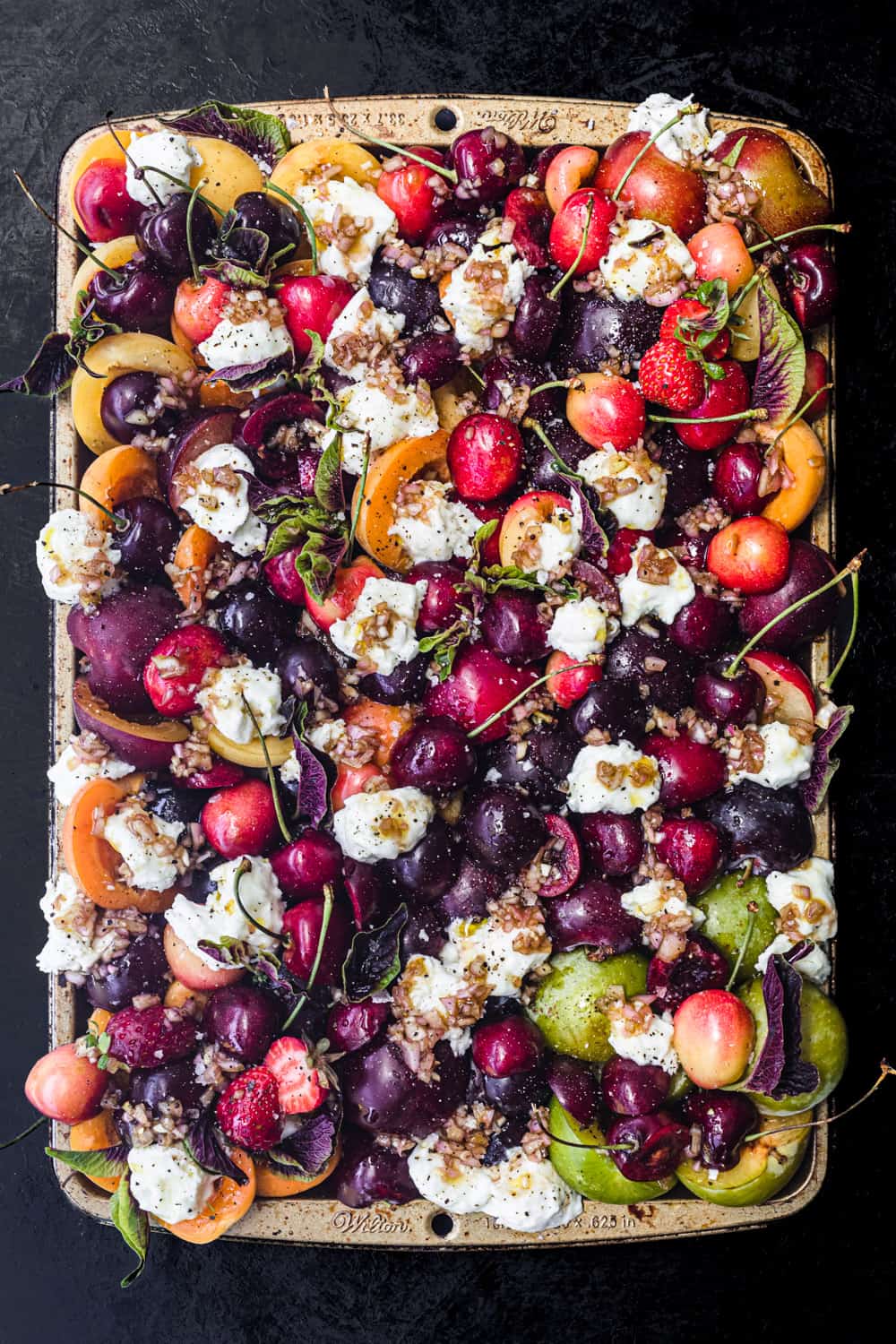 a very colorful peaches and other stone fruits and burrata salad; arranged on a tray on a dark background; overhead shot.