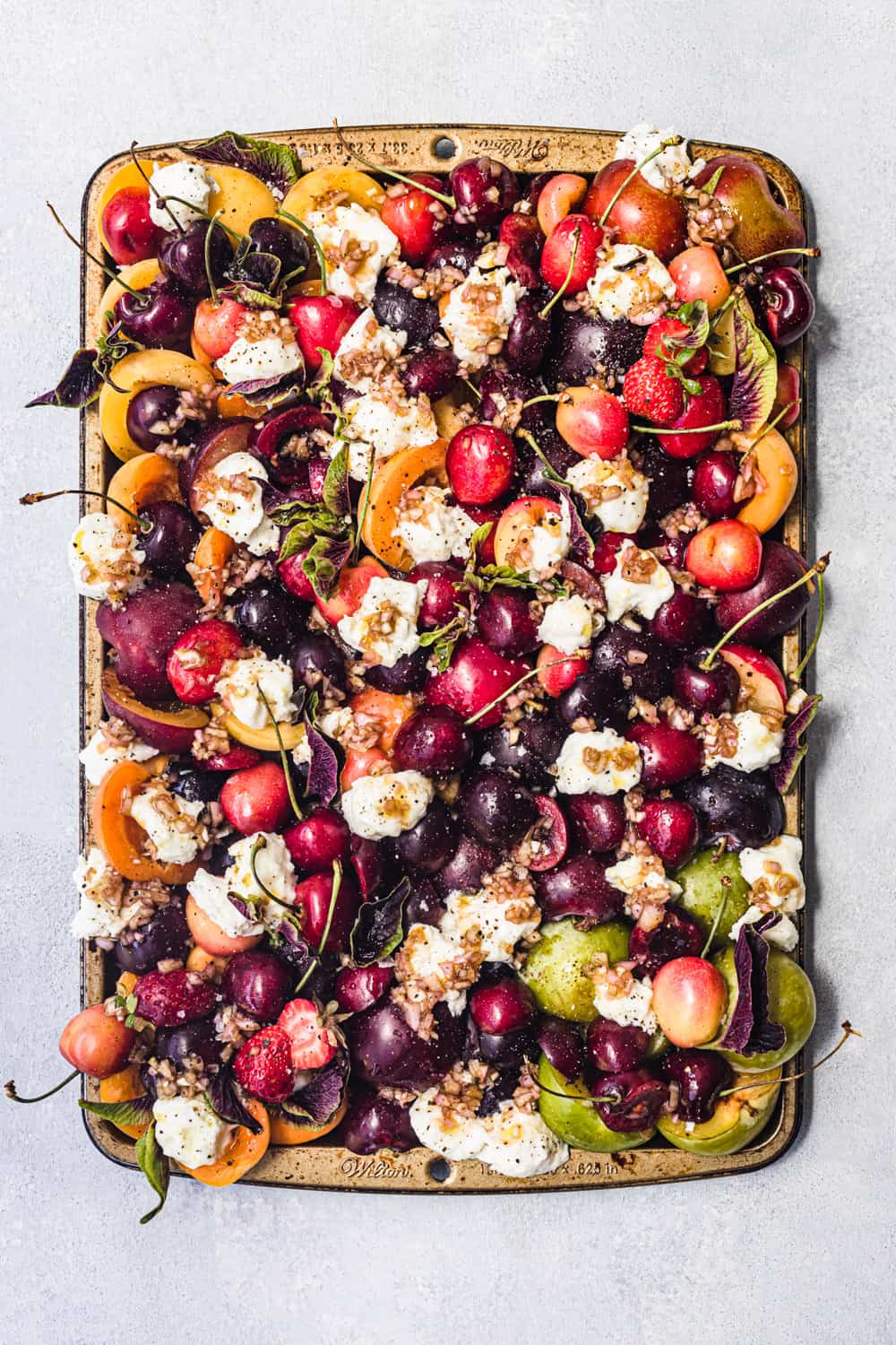 peaches, cherries, plums, apricots and nectarines with burrata, basil and vinaigrette arranged on a tray, overhead shot on a light background.