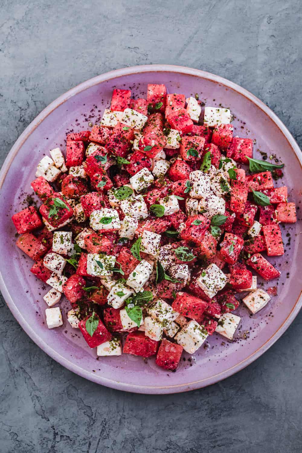 Watermelon feta salad with fresh herbs and spices. Overhead shot, on a pink plate and grey background.