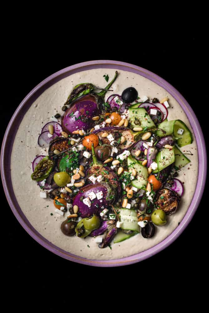 hummus is on a pink plate, with seasonal fruits and veggies, overhead on a black background.