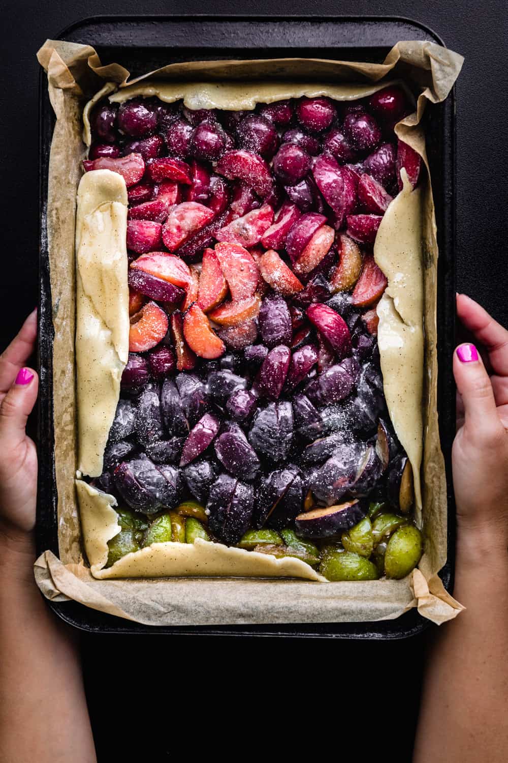 Daniela Gerson holding a very colorful cherry and plum galette; overhead on a dark background.