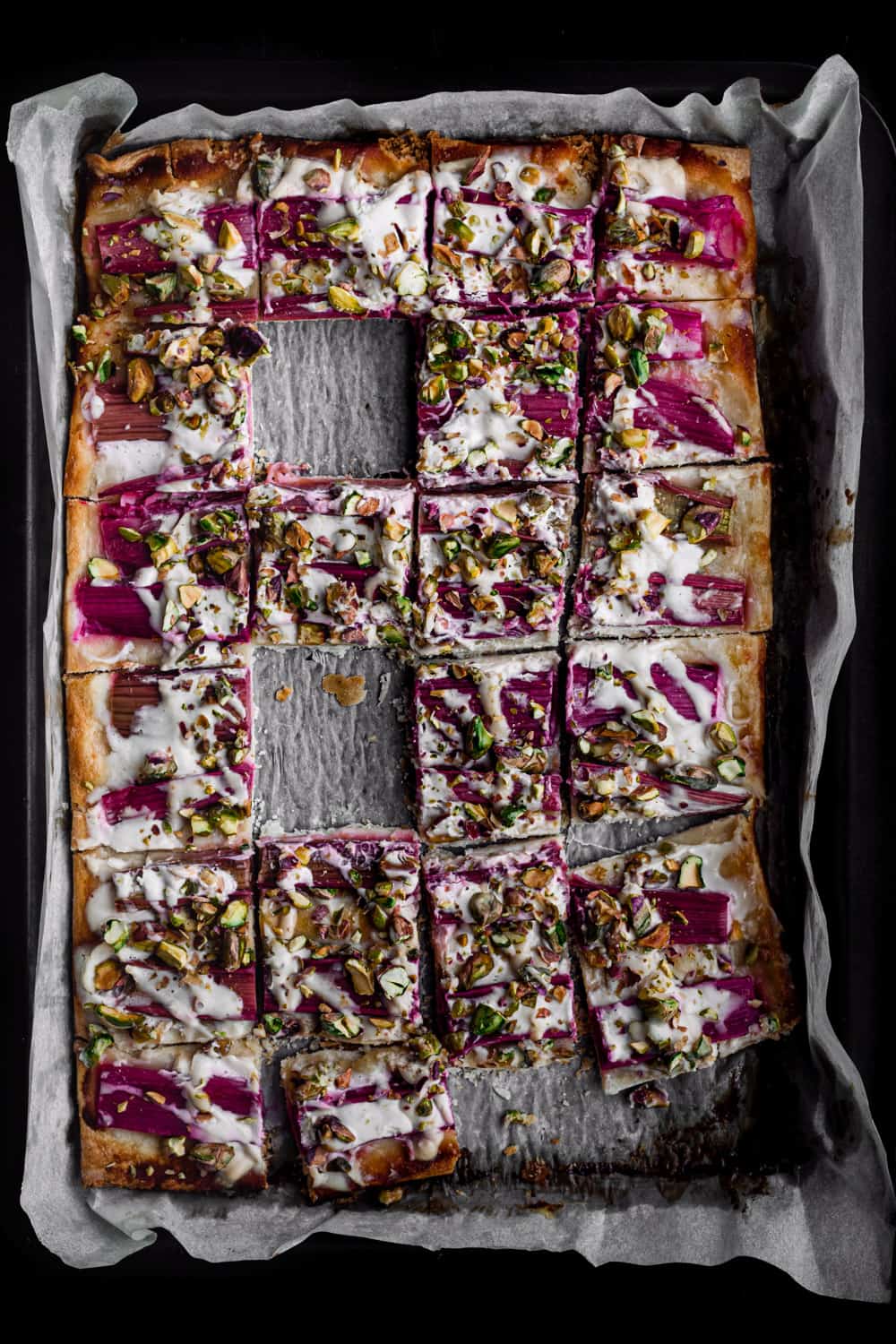 Rhubarb tart sprinkled with pistachios and drizzled with honey, cut into square, overhead and a few squares missing.