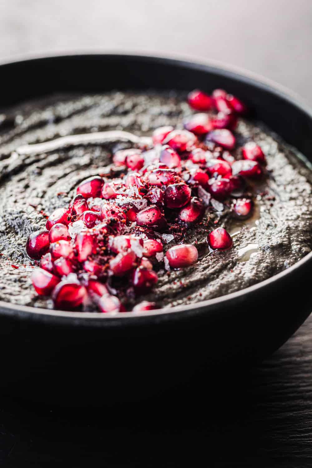 black hummus topped with pomegranate seeds in a black bowl, side angle