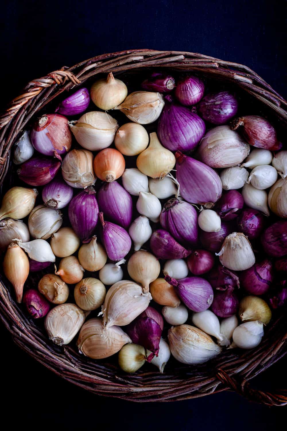 Raw purple shallots and little white and yellow onions in a basket on a dark background.