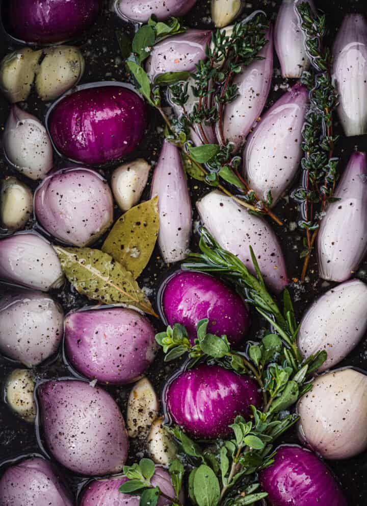 raw shallots submerged in olive oil with garlic, bay leaves, fresh herbs, salt and pepper; overhead shot.