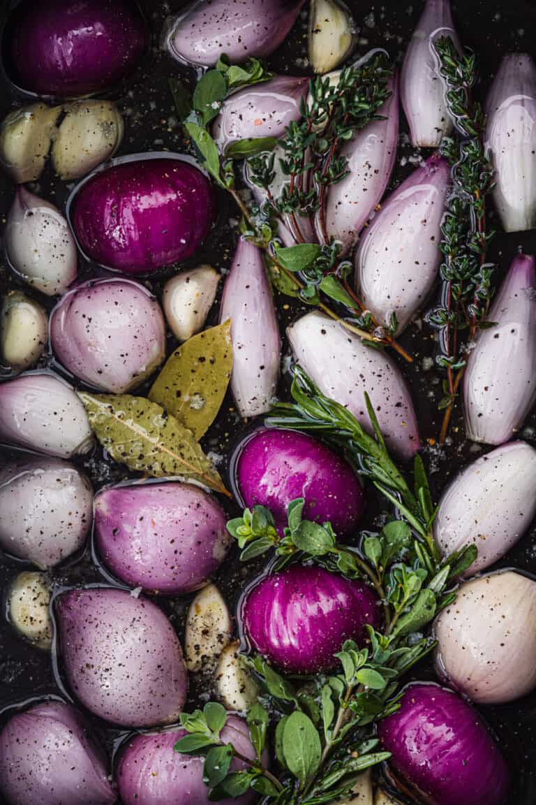 raw shallots submerged in olive oil with garlic, bay leaves, fresh herbs, salt and pepper; overhead shot.