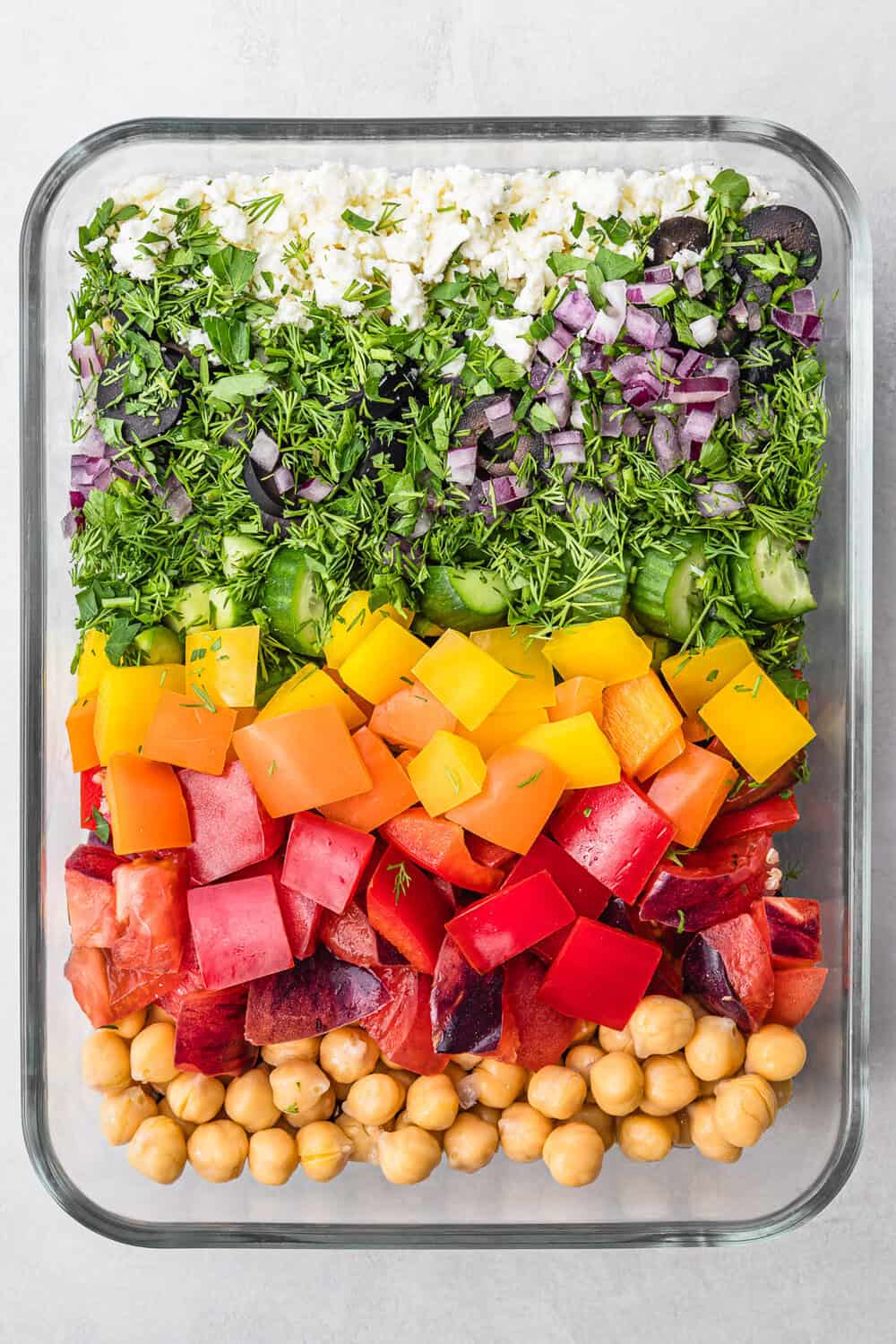 Overhead shot of colorful salad in a glass rectangular dish – green herbs, purple red onion, orange ellow bell peppers, juicy red tomatoes, and golden chickpeas on a white background.
