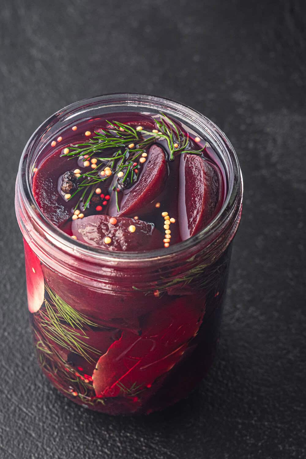Quick pickled beets in a glass jar with fresh dill, mustard seeds, bay leaves, and peppercorns visible at the top and in the jar, on a dark background.