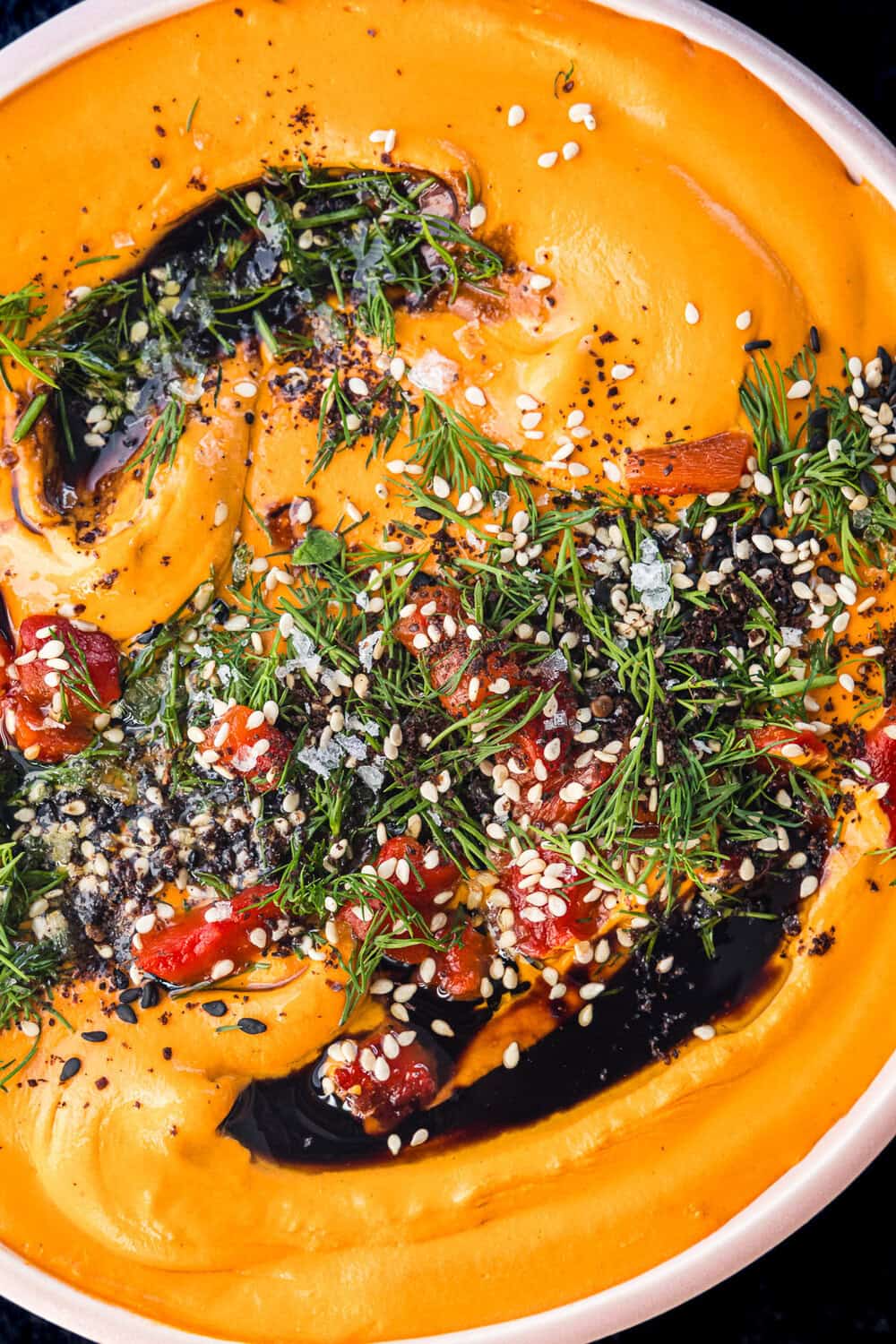 Roasted red pepper hummus in a pink bowl with lots of toppings like fresh dill, black and white sesame seeds, flaky sea salt and a drizzle of olive oil, and on a black background.
