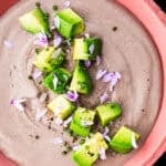 Overhead shot of creamy black bean dip topped with avocado, chives, purple chive flowers, and flaky salt in a pink ceramic bowl on a black background.