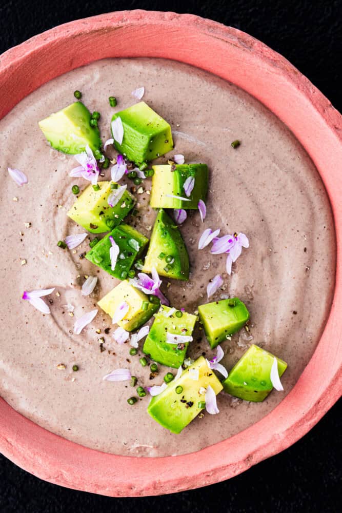Overhead shot of creamy black bean dip topped with avocado, chives, purple chive flowers, and flaky salt in a pink ceramic bowl on a black background.