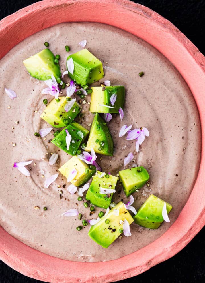 Overhead shot of creamy black bean dip topped with avocado, chives, purple chive flowers, and flaky salt in a pink ceramic bowl on a black background.
