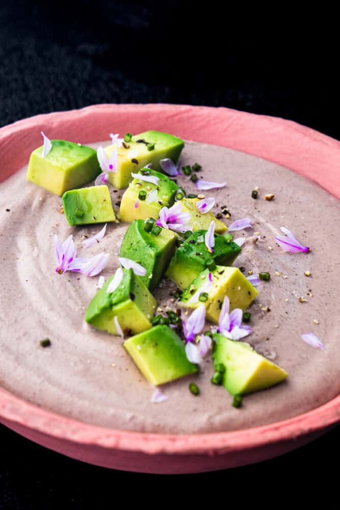 Side angle close-up of creamy black bean dip topped with avocado, chives, purple chive flowers, and flaky salt in a pink ceramic bowl on a black background.