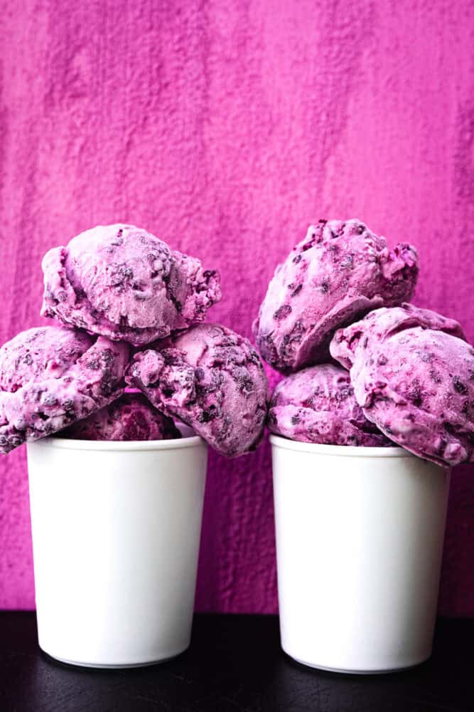 Blackberry cheesecake ice cream scoops piled high in two white cups against a hot pink textured background.