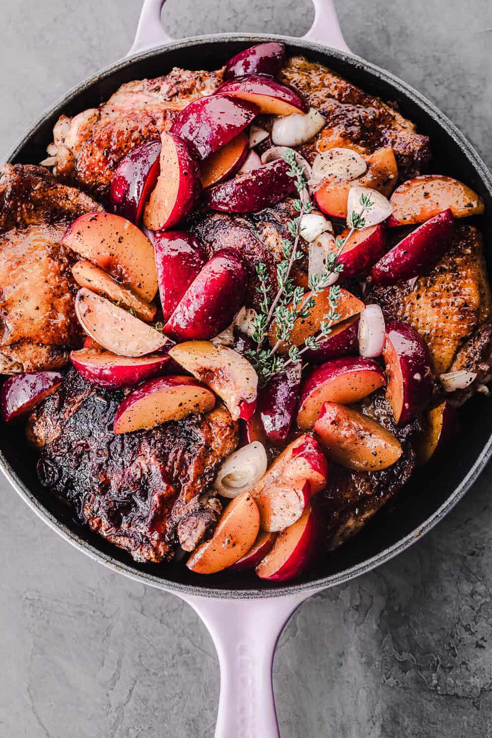 Crispy golden-brown chicken thighs with, caramelized peaches, shallots and thyme in a lavender cast iron skillet on a grey stone background, overhead shot.