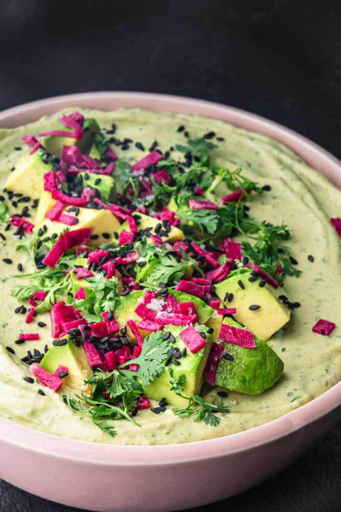 Avocado hummus in a pink bowl topped with cubed avocado, magenta pickled radish, black sesame seeds, and fresh cilantro, on a black background, side angle shot.