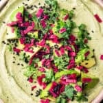 Overhead shot of avocado hummus in a pink bowl topped with cubed avocado, magenta pickled radish, black sesame seeds, and fresh cilantro, on a black background.