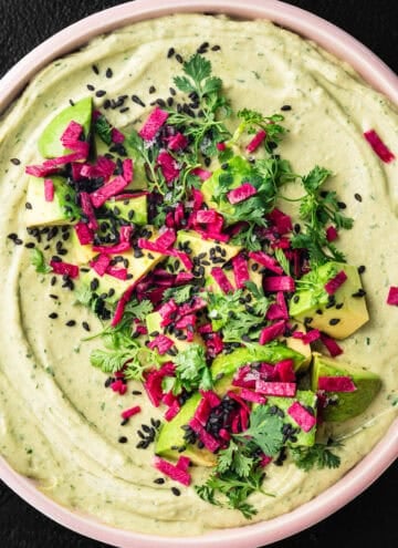 Overhead shot of avocado hummus in a pink bowl topped with cubed avocado, magenta pickled radish, black sesame seeds, and fresh cilantro, on a black background.