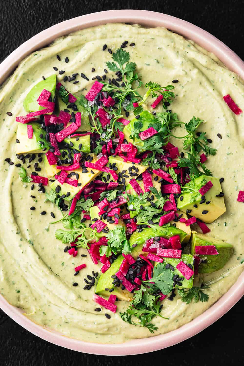 Overhead shot of avocado hummus in a pink bowl topped with cubed avocado, magenta pickled radish, black sesame seeds, and fresh cilantro, on a black background.