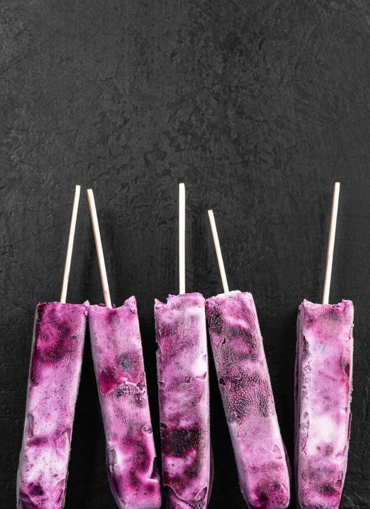 Blueberry yogurt popsicles, with purple and white swirls, standing upright against a dark grey textured background