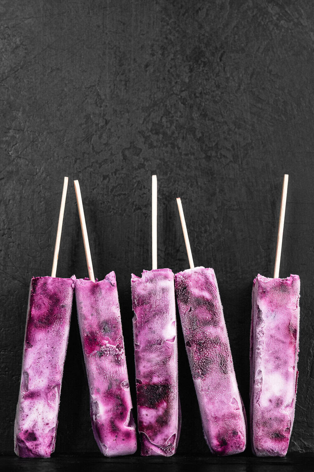 Blueberry yogurt popsicles, with purple and white swirls, standing upright against a dark grey textured background