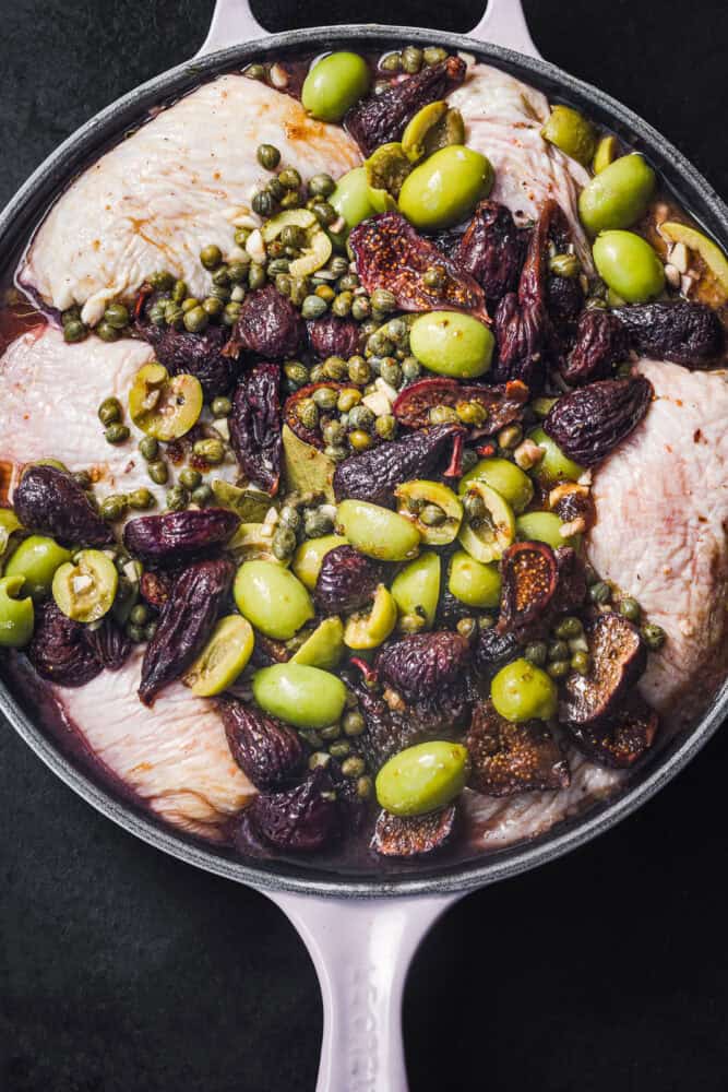 Overhead close-up of raw chicken thighs marinating in a lavender skillet with dried figs, green olives, capers, and garlic, against a dark background.
