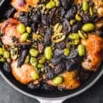 Overhead shot of roasted golden-brown chicken thighs in a skillet topped with green olives, dried figs, capers, and a whole roasted head of garlic, on a dark background.