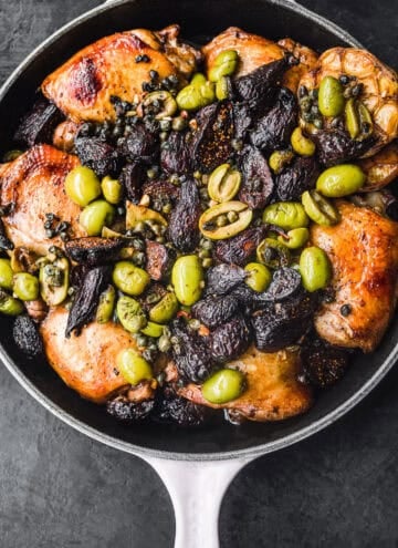 Overhead shot of roasted golden-brown chicken thighs in a skillet topped with green olives, dried figs, capers, and a whole roasted head of garlic, on a dark background.