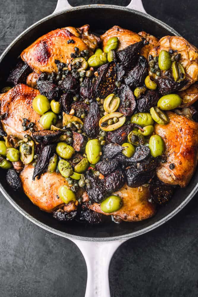 Overhead shot of roasted golden-brown chicken thighs in a skillet topped with green olives, dried figs, capers, and a whole roasted head of garlic, on a dark background.