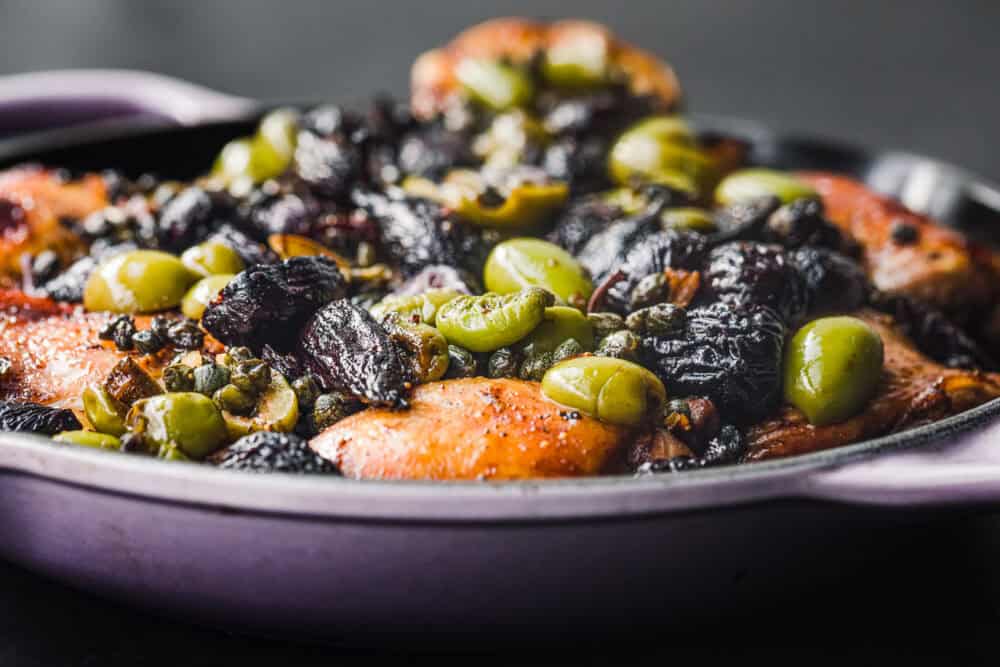 Side angle close-up of Chicken Marbella with dried figs, green olives, and capers in a lavender skillet on a dark background.