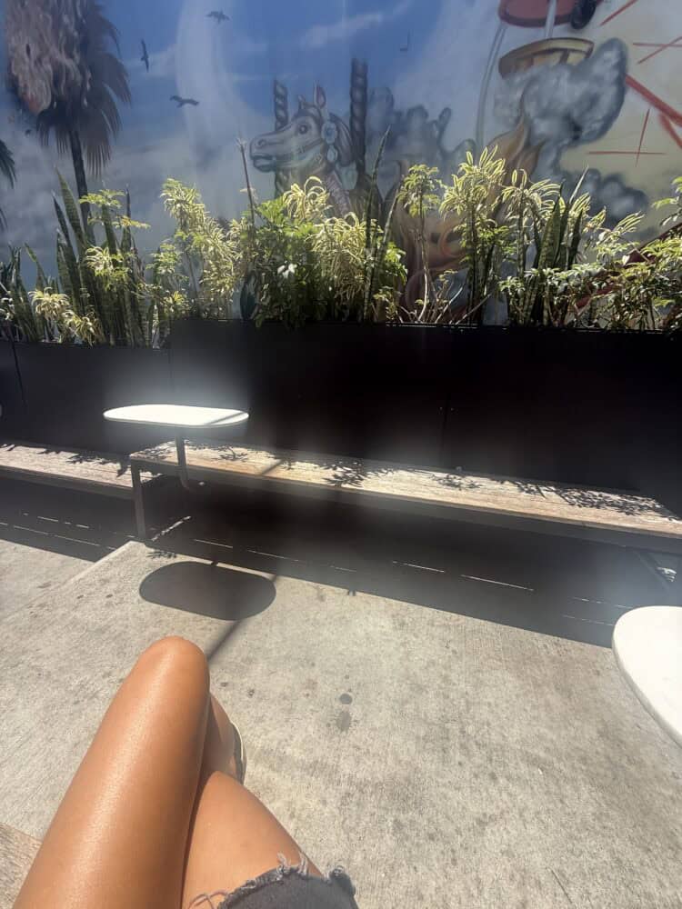 Legs stretched out on an outdoor bench at Intelligentsia Coffee on Abbot Kinney, with potted plants, a mural, and a small round table in the background on a sunny day.