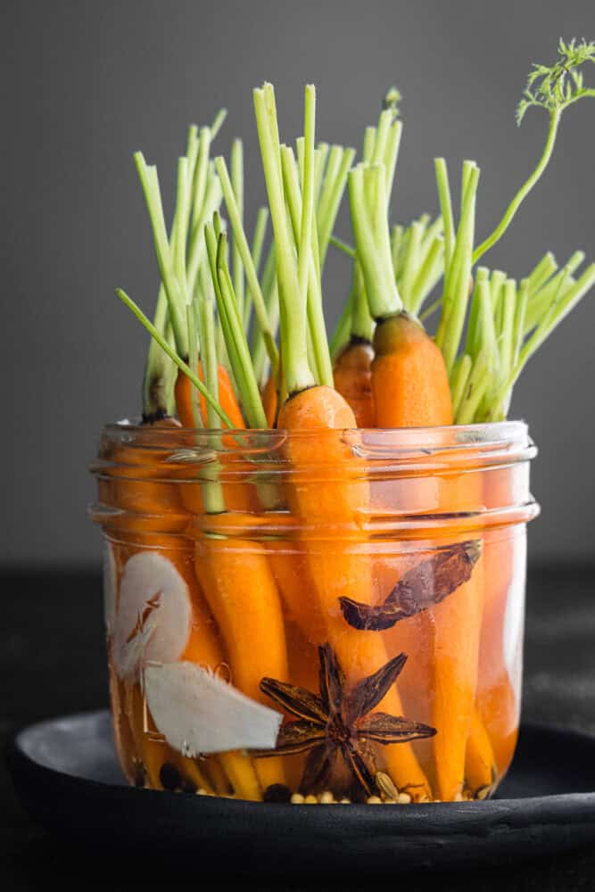 Pickled carrots with green tops pickled in a glass jar with sliced garlic, fennel seeds, mustard seeds and cumin seeds on a dark background, close-up side angle