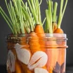 Pickled carrots with green tops in a glass jar with sliced garlic, star anise, mustard seeds, cumin seeds, and fennel seeds, on dark grey background, close-up side angle