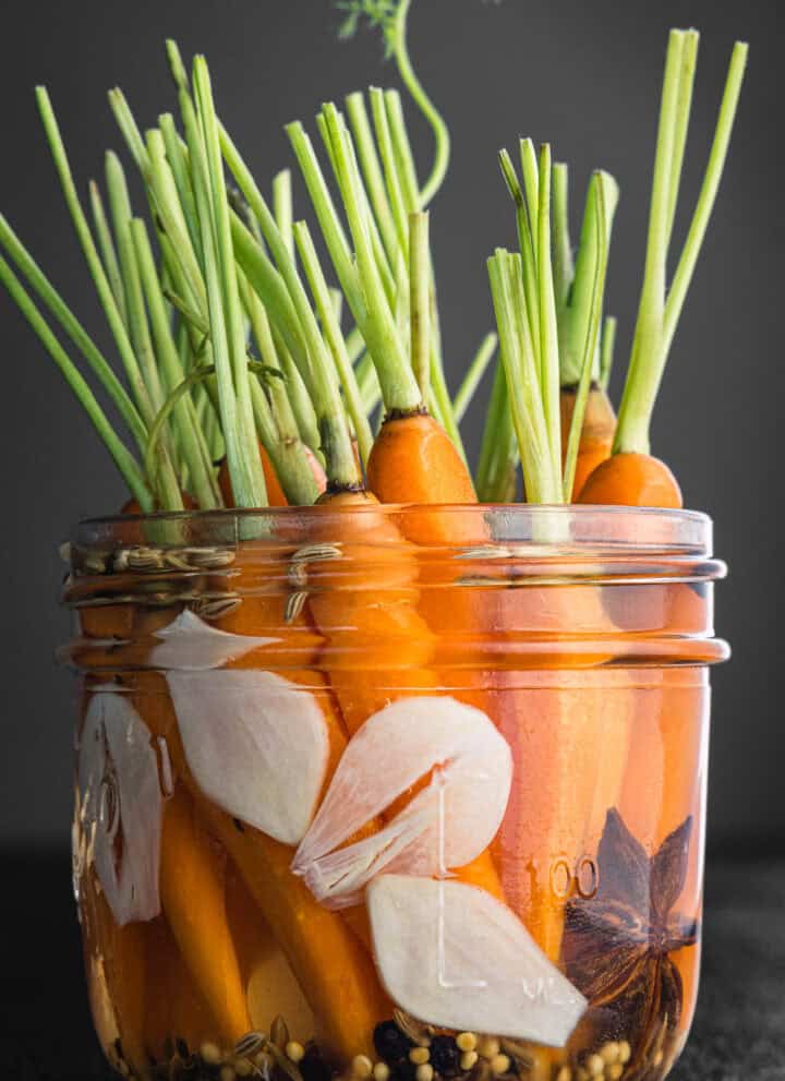 Pickled carrots with green tops in a glass jar with sliced garlic, star anise, mustard seeds, cumin seeds, and fennel seeds, on dark grey background, close-up side angle