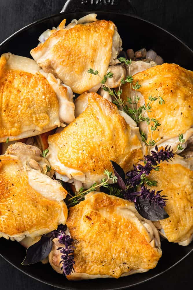 Seared bone-in, skin-on chicken thighs with golden crispy skin, garnished with fresh thyme and purple basil, overhead in a black cast iron skillet on a dark background.