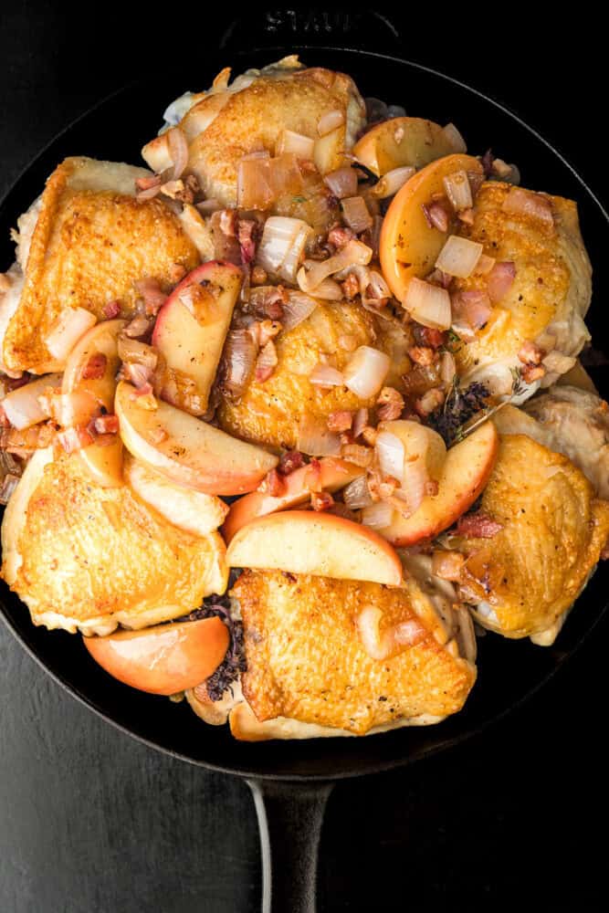 Roasted bone-in chicken thighs topped with apple wedges, chopped shallots, and diced pancetta, overhead in a black cast iron skillet on a dark background.